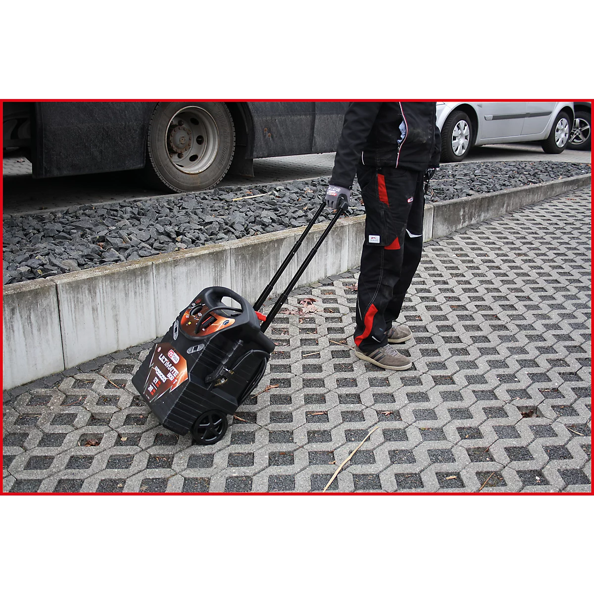 Person zieht eine schwarze Trolley-Box mit Rädern über ein Pflaster. Im Hintergrund ein Bus und ein Auto.